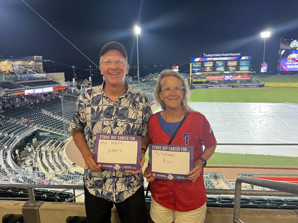 Ron and his wife Nancy hold signs at a baseball stadium. Ron's says "Strike Out Cancer For...My Wife, Nancy" and Nancy's says "Strike Out Cancer For...My husband, Ron"